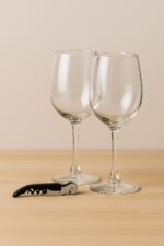 Dos copas de vino con abridor metálico sobre superficie de madera clara y fondo beige, set de regalo corporativo MQM.